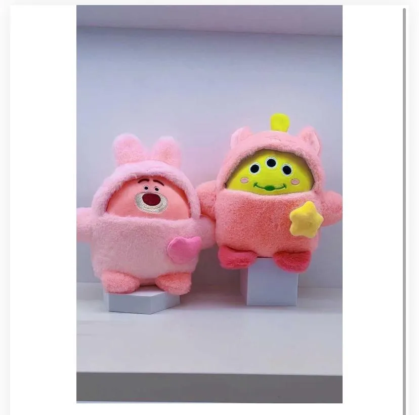 A round, pink plush character dressed in a 781657soft, fuzzy pink hooded outfit with ears on top. It has a central embroidered facial feature resembling a strawberry bear